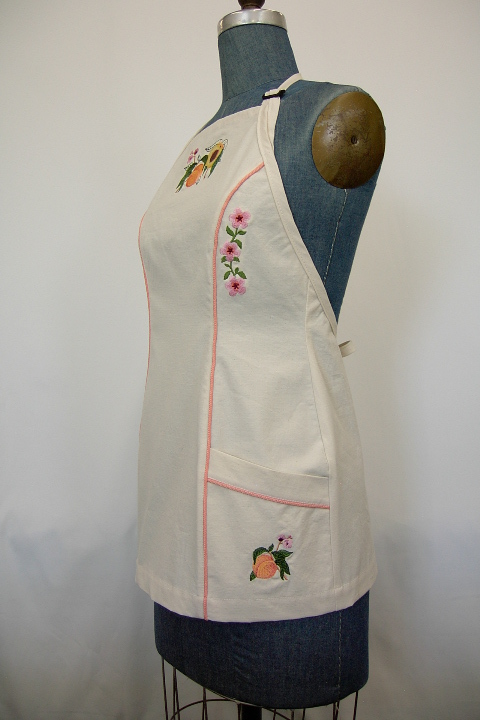 Women's Bib apron style W740 shortened 5 inches; Shown in Natural, 100% cotton riptstop with two side hip tailored welt pockets, bisque piping on pocket welts & along princess seams, embroidered peach botanical on center chest, embroidered peach blossom chains; one along each princess seam & individual peaches; one on each pocket.