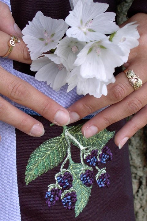 Close up image of blackberry embroidery on the pocket of Women's Chef Coat style BSW104; Shown in Lavender & black, 100% cotton petti point pique & 100% cotton Gabardine, two front hip tailored welt pockets, Copen piping (collar, front, cuffs & pockets), Black Gabardine, hand tied knot buttons & blackberry embroideries on the chests, pockets, & back of collar.