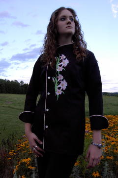 Women's Chef Coat Style CBW105: Shown in Black 100% cotton Supima gabardine, with a left sleeve tailored welt pocket, Pink Sham piping (cuffs, collar, front), faux Mother-of-Pearl buttons & embroidered Easter Lily clusters.